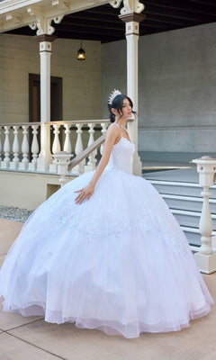 A young woman stands to the side in front of a porch with white railings and steps, wearing the Juliet JT1496HW Long Wedding Dress—a white tulle ball gown adorned with delicate floral lace appliques—paired with a sparkling tiara.