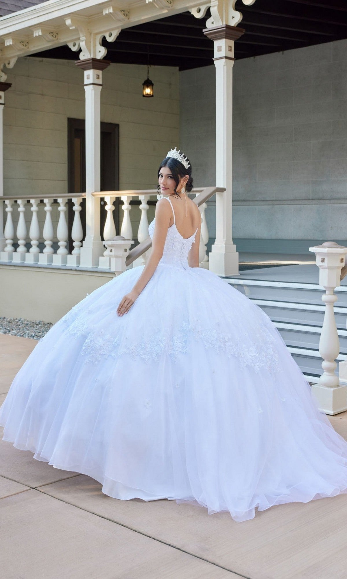 A woman wearing the Juliet JT1496HW Long Wedding Dress, featuring elegant floral lace appliques, and a sparkling tiara stands outside a building with white railings, glancing over her shoulder.