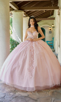 A young woman stands on a stone porch beneath a wooden roof, hands clasped in front, wearing the Quinceanera Dress JT1497J By Juliet in blush with delicate floral lace appliques and a sparkling tiara.