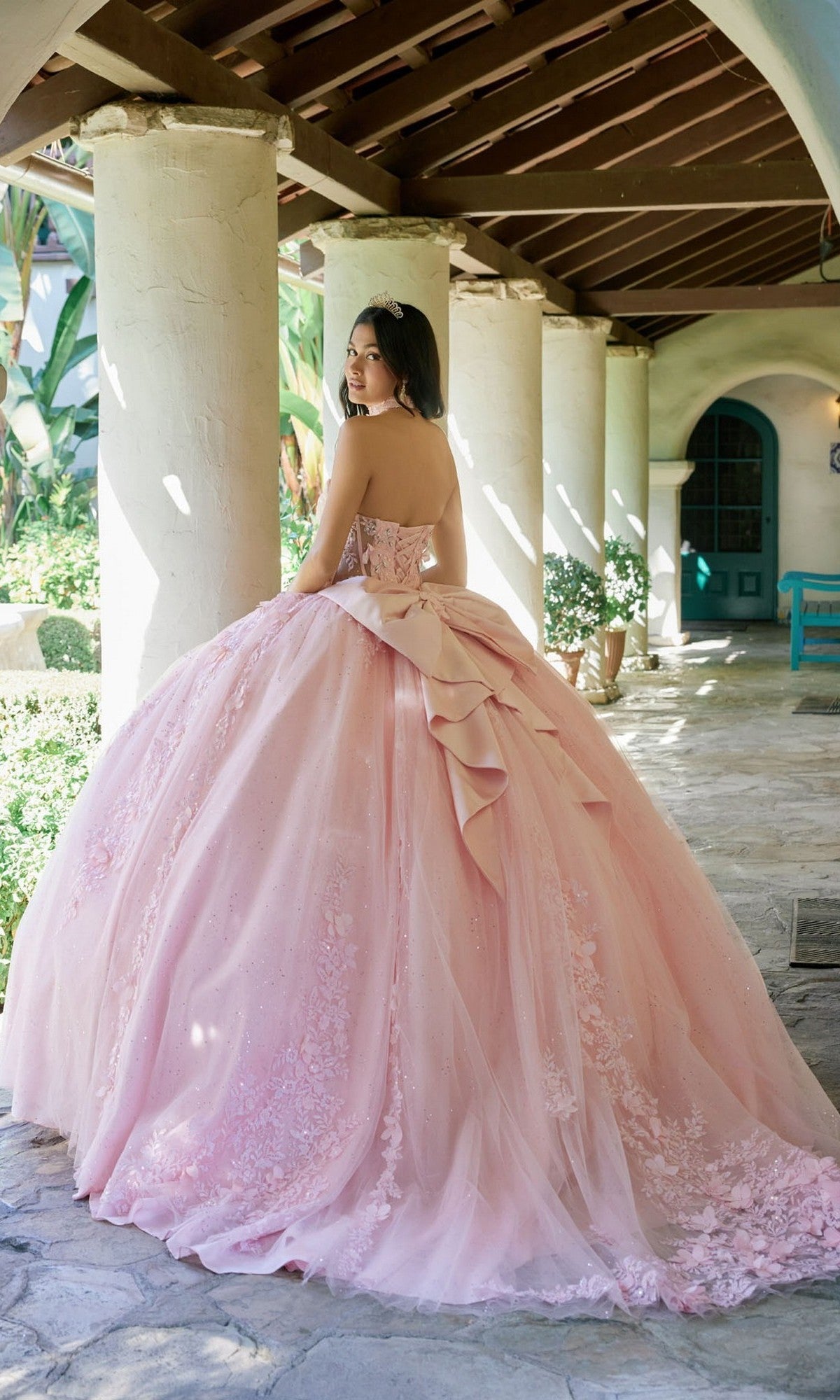 A woman stands in a stone hallway wearing the Quinceanera Dress JT1497J By Juliet—a strapless pastel-pink ballgown adorned with a large bow and delicate floral lace appliques on the skirt.