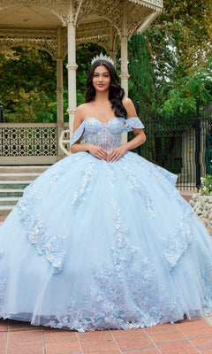 A young woman wearing the Quinceanera Dress JT1498J By Juliet in Bahama Blue, featuring an off-the-shoulder design with a flowing ballgown skirt and a tiara, stands outdoors in front of a gazebo.