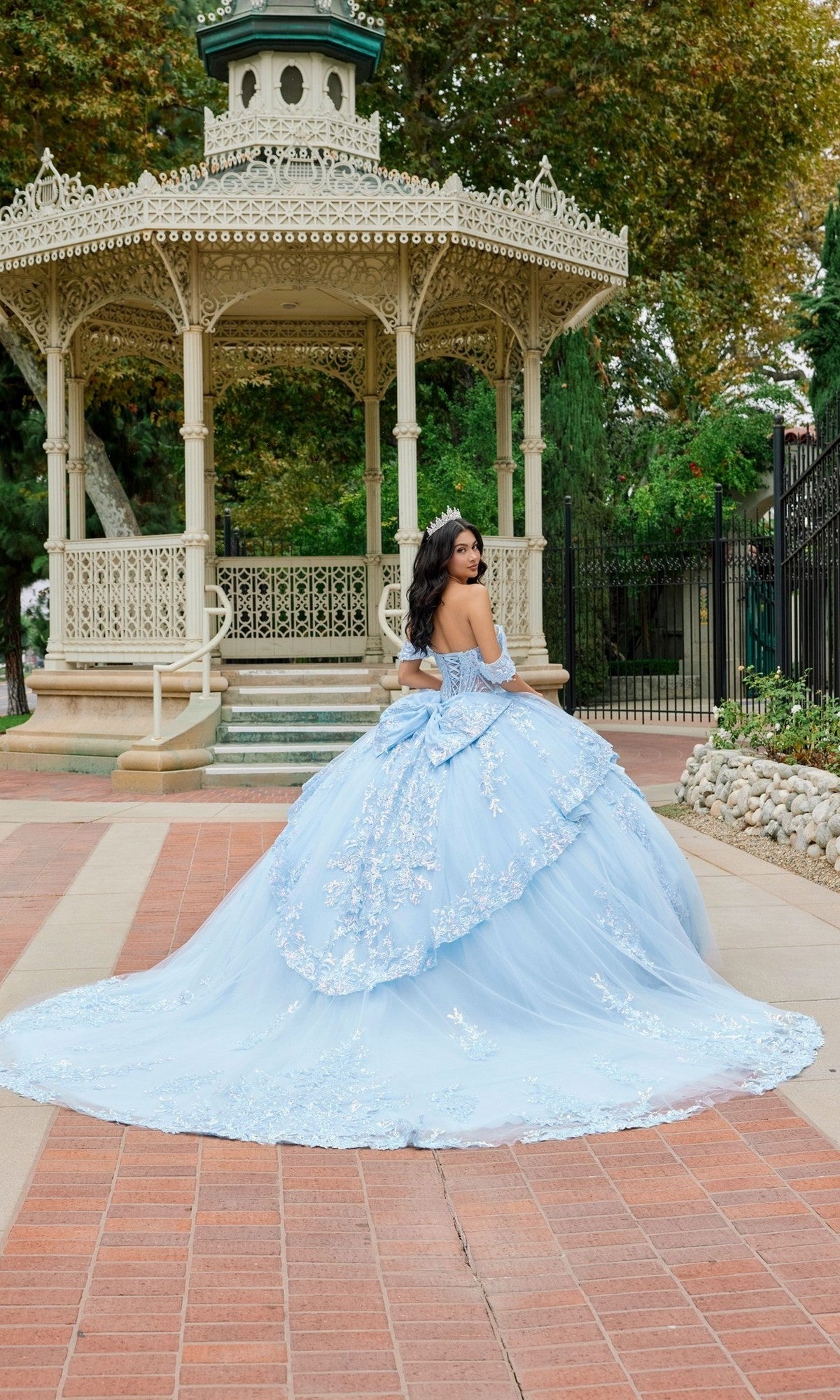 Wearing the Quinceanera Dress JT1498J by Juliet, a young woman stands in front of an ornate gazebo, gazing over her shoulder. The light blue off-the-shoulder ballgown skirt flows elegantly as she is framed by trees and lush greenery in the background.