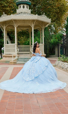 Wearing the Quinceanera Dress JT1498J by Juliet, a young woman stands in front of an ornate gazebo, gazing over her shoulder. The light blue off-the-shoulder ballgown skirt flows elegantly as she is framed by trees and lush greenery in the background.