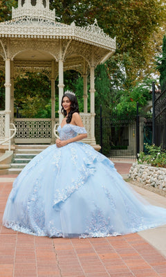 A young woman wearing the Quinceanera Dress JT1498J By Juliet—a light blue, embroidered off-the-shoulder gown with a flowing ballgown skirt and a tiara—stands in front of a decorative gazebo in a garden setting.