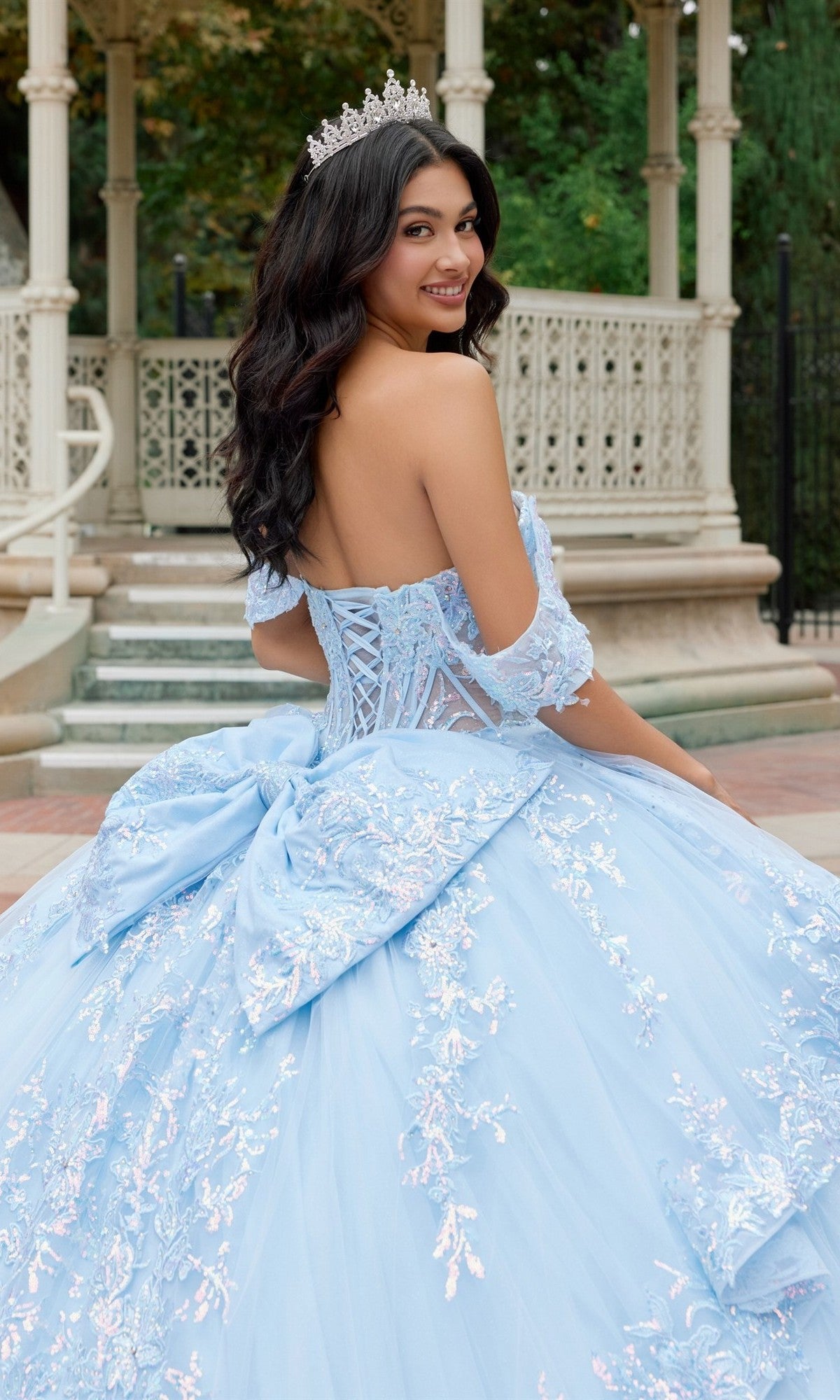 A young woman wearing the Quinceanera Dress JT1498J By Juliet—a light blue, off-the-shoulder gown with lace details and a large bow—adorns a tiara as she smiles over her shoulder in front of an ornate outdoor gazebo.