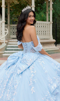 A young woman wearing the Quinceanera Dress JT1498J By Juliet—a light blue, off-the-shoulder gown with lace details and a large bow—adorns a tiara as she smiles over her shoulder in front of an ornate outdoor gazebo.