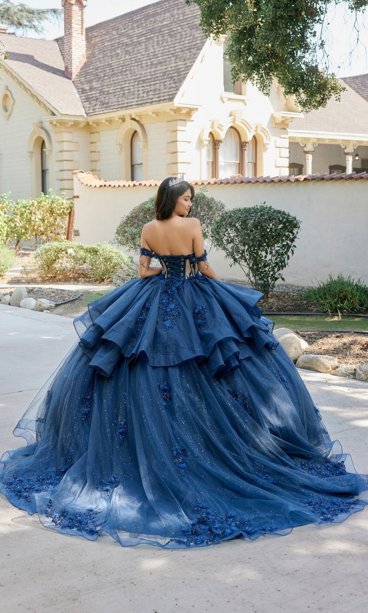 Wearing the Quinceanera Dress JT1501J By Juliet—an off-the-shoulder, navy-blue ballgown featuring a beaded corset bodice and a glittery skirt adorned with floral details—a woman stands on a path outside a cream-colored building.