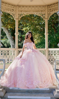 A woman wearing the Quinceanera Dress JT1503J By Juliet in blush, featuring a delicate corset bodice and lace details, stands on steps in a decorative outdoor gazebo surrounded by greenery.