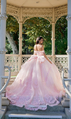 Wearing the Quinceanera Dress JT1503J By Juliet, a woman stands on steps before an ornate gazebo, her off-shoulder pastel-pink gown with a corset bodice flowing gracefully as she faces away amid lush greenery and sunlight.