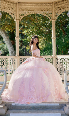 A young woman wearing the Quinceanera Dress JT1503J By Juliet, featuring a graceful corset bodice in pastel pink, stands under an ornate gazebo in a garden and looks over her shoulder toward the camera.