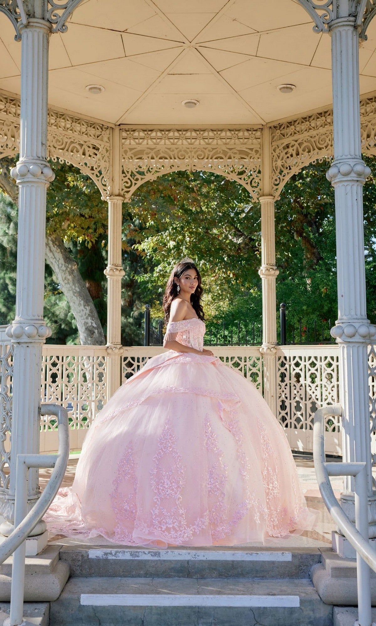 A young woman stands under an ornate gazebo, sunlight filtering through the greenery behind her, wearing the Quinceanera Dress JT1503J By Juliet—a pastel-pink gown featuring a tulle ballgown skirt and corset bodice.