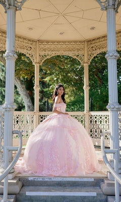 A young woman stands under an ornate gazebo, sunlight filtering through the greenery behind her, wearing the Quinceanera Dress JT1503J By Juliet—a pastel-pink gown featuring a tulle ballgown skirt and corset bodice.