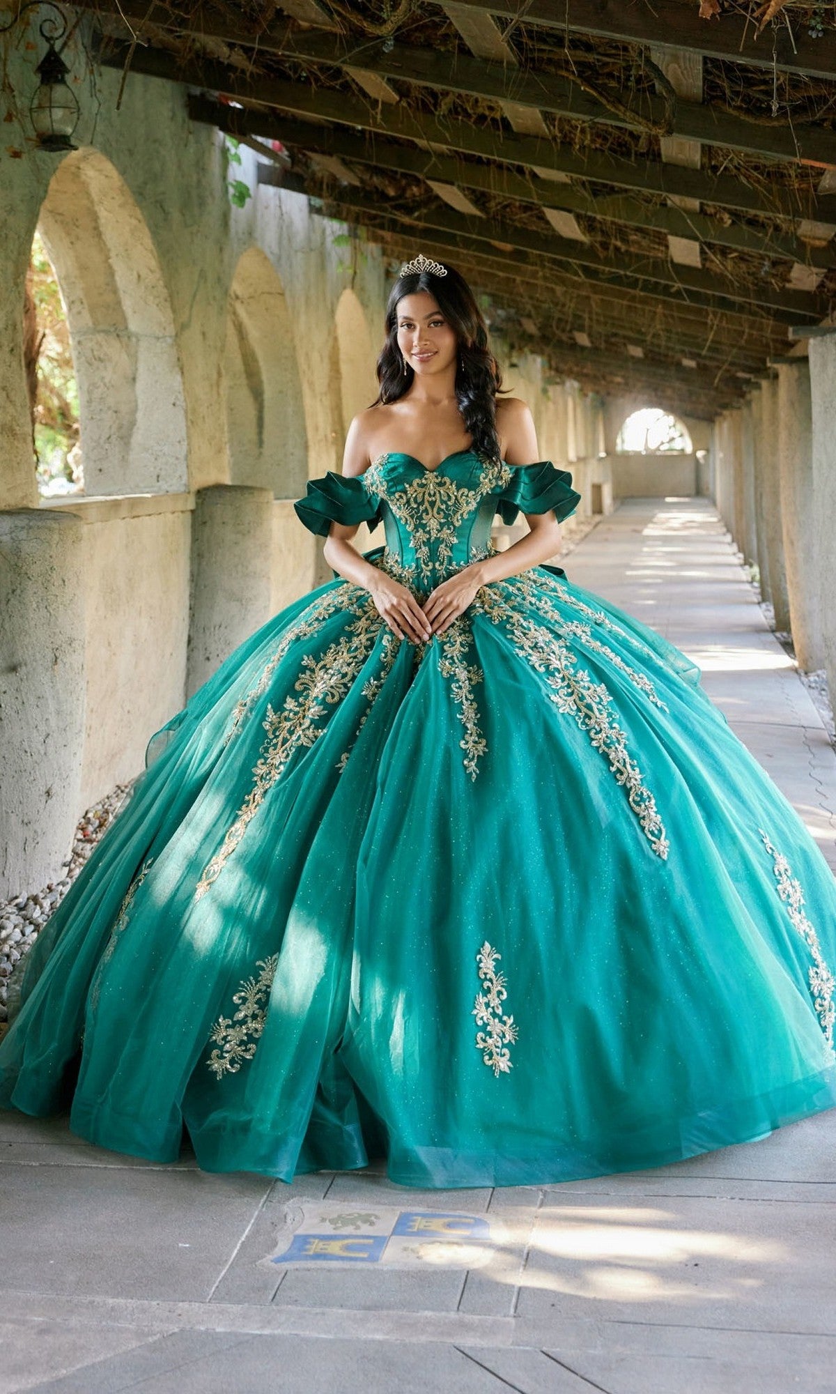 A woman stands in a stone archway corridor, holding her hands together at her waist and wearing a tiara. She is dressed in the Quinceanera Dress JT1504J By Juliet in Green/Gold, which features gold embroidery, a glitter tulle skirt, and a corset bodice.