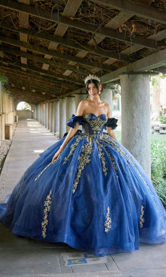 A woman stands gracefully under a covered walkway, wearing the Quinceanera Dress JT1504J By Juliet in Navy/Gold—a stunning ball gown featuring ornate gold embroidery, a glitter tulle skirt, and a sparkling tiara.