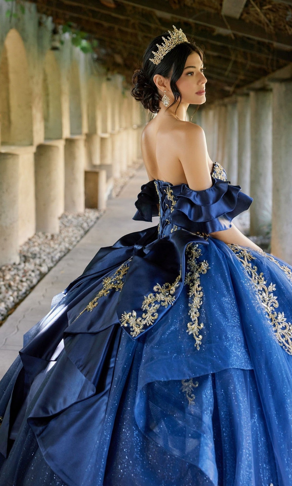 A woman wearing the Quinceanera Dress JT1504J By Juliet, an ornate blue ball gown with a corset bodice and tiara, stands outdoors under a covered walkway, facing away as she looks over her shoulder.