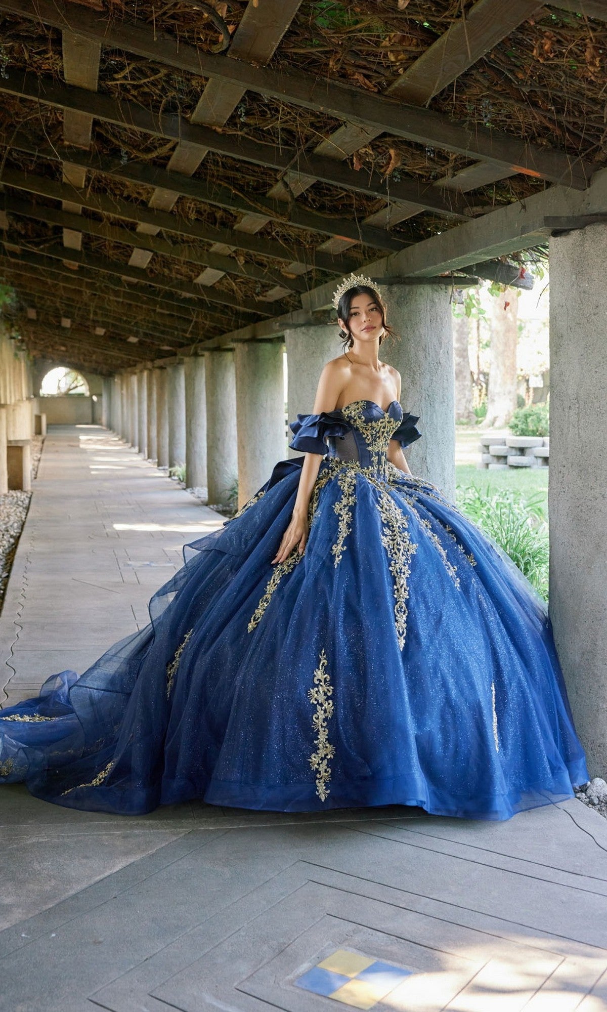 A woman wearing the Quinceanera Dress JT1504J By Juliet, a blue ball gown featuring gold embroidery and a fitted corset bodice, stands under a pergola adorned with stone columns and greenery overhead.