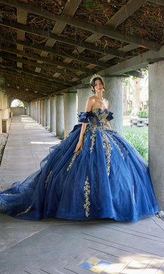 A woman wearing the Quinceanera Dress JT1504J By Juliet, a blue ball gown featuring gold embroidery and a fitted corset bodice, stands under a pergola adorned with stone columns and greenery overhead.