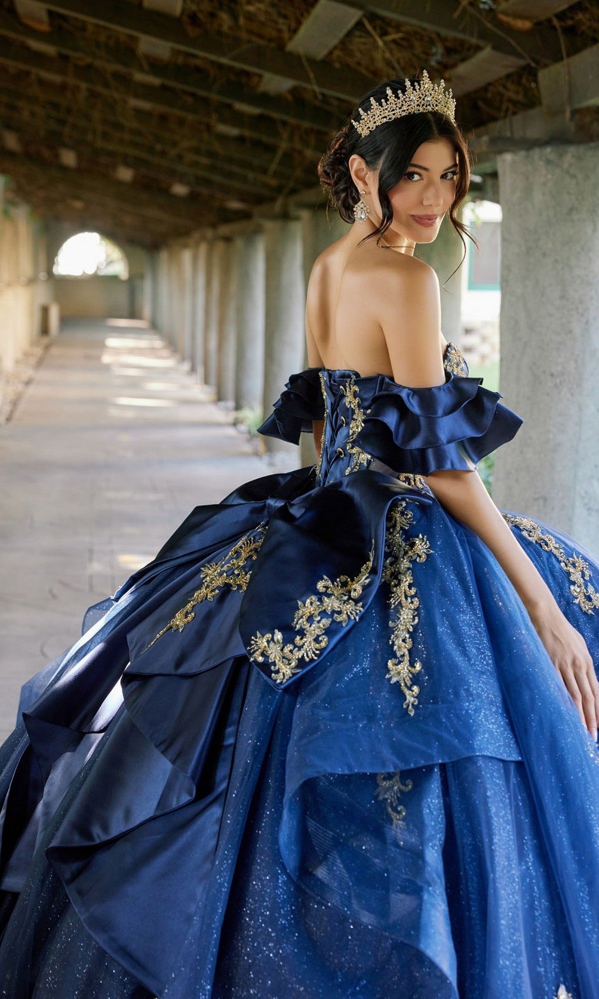 Wearing the Quinceanera Dress JT1504J By Juliet—a blue ball gown adorned with gold embroidery, a corset bodice, and a glitter tulle skirt—a woman stands in a corridor, gazing over her shoulder as her gold tiara shimmers in the light.