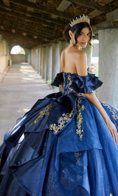 Wearing the Quinceanera Dress JT1504J By Juliet—a blue ball gown adorned with gold embroidery, a corset bodice, and a glitter tulle skirt—a woman stands in a corridor, gazing over her shoulder as her gold tiara shimmers in the light.