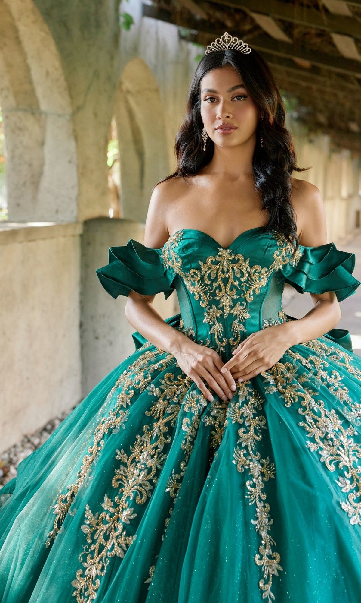A woman stands in a stone corridor with her hands clasped in front of her, wearing the Quinceanera Dress JT1504J By Juliet—an elegant green and gold off-shoulder ball gown featuring a glitter tulle skirt and corset bodice—topped with a sparkling tiara.