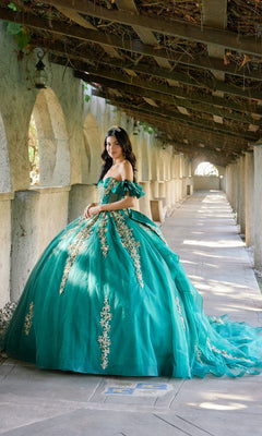 A young woman in the Quinceanera Dress JT1504J By Juliet, a teal ball gown adorned with gold embroidery, stands in a sunlit arched walkway, looking at the camera. The elegant dress features a glitter tulle skirt and a fitted corset bodice, adding sophistication to the radiant scene.