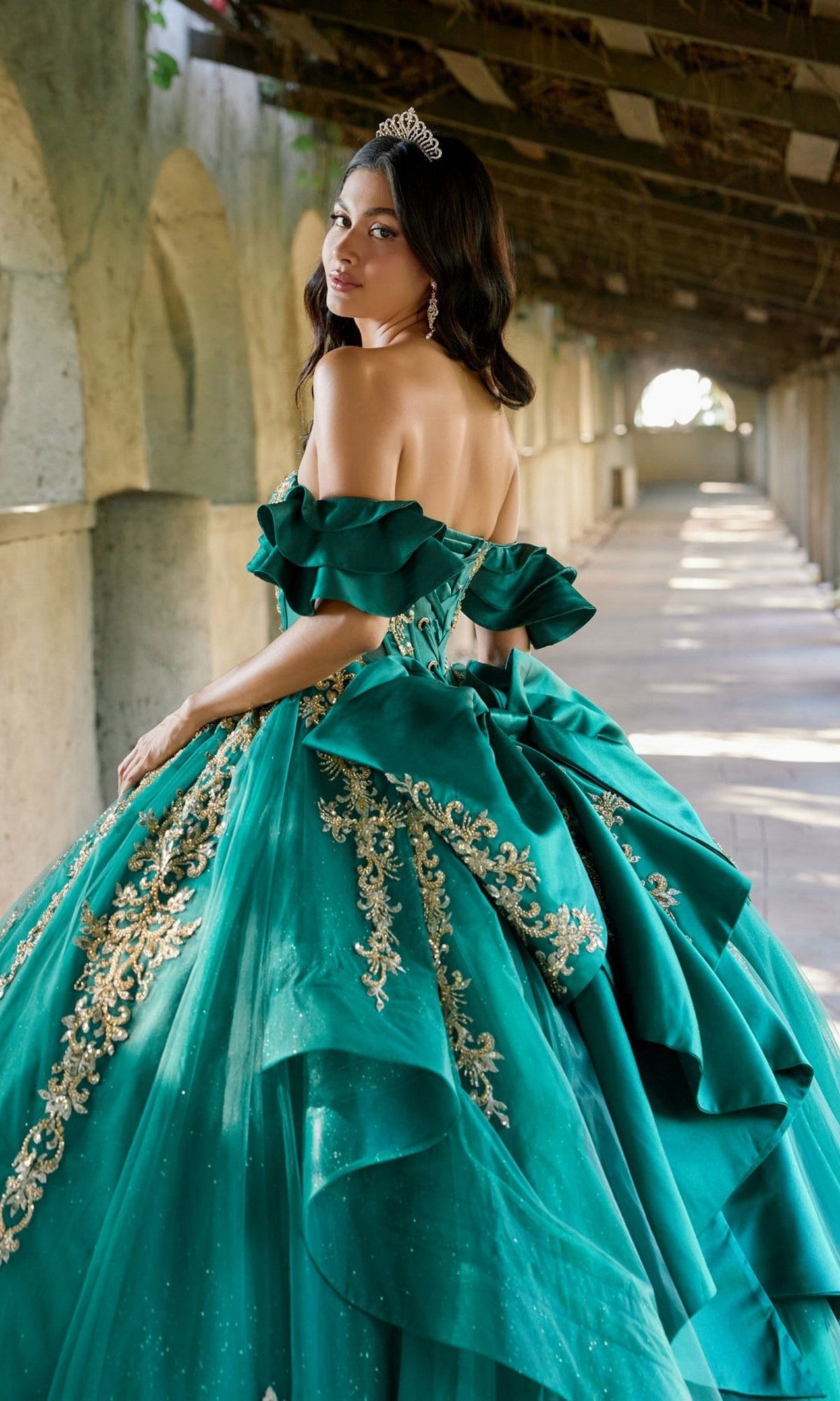 A woman in the Quinceanera Dress JT1504J By Juliet—a green, off-shoulder ball gown featuring a glitter tulle skirt and corset bodice—stands in a sunlit, arched corridor, looking back over her shoulder as her tiara catches the light.