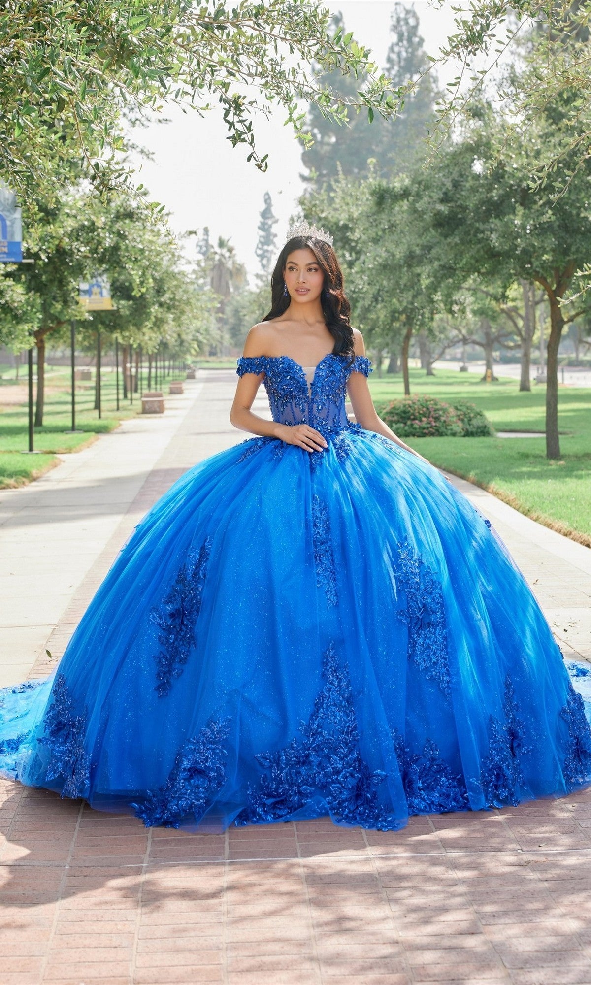 A young woman stands on a tree-lined walkway in a park, looking at the camera while wearing the Quinceanera Dress JT1505J By Juliet in Royal Blue and a sparkling tiara.