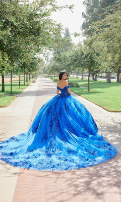 A woman wearing the Quinceanera Dress JT1505J by Juliet, a vibrant blue ball gown with a sweeping train, stands on a tree-lined path in a park, facing away from the camera.