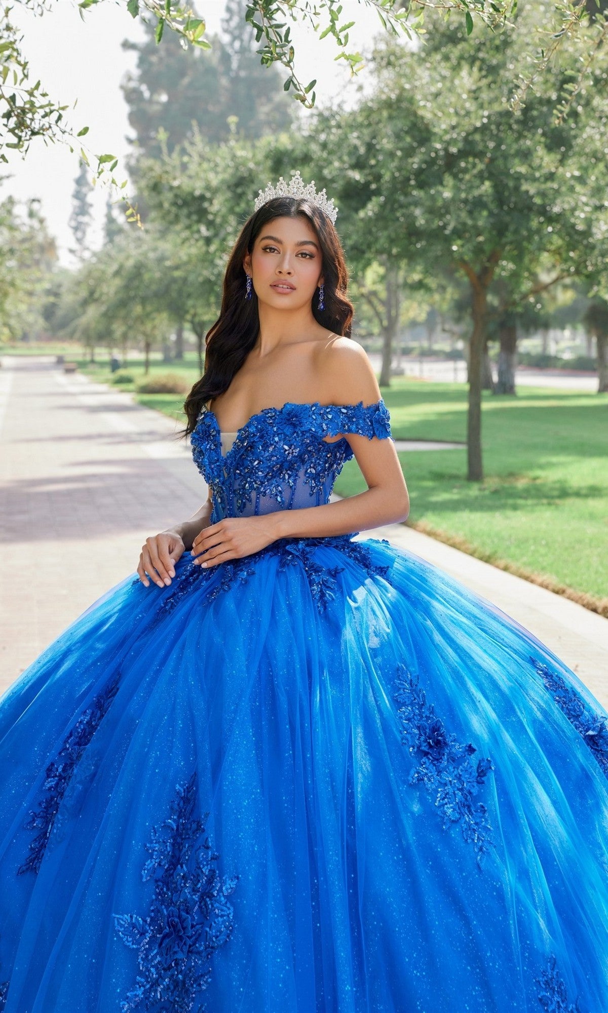 Wearing the Quinceanera Dress JT1505J by Juliet, a woman in a blue ball gown and tiara stands on a tree-lined path in a park, looking at the camera. The elegant off-the-shoulder design of the dress accentuates her graceful pose, lending an air of timeless beauty to the scene.