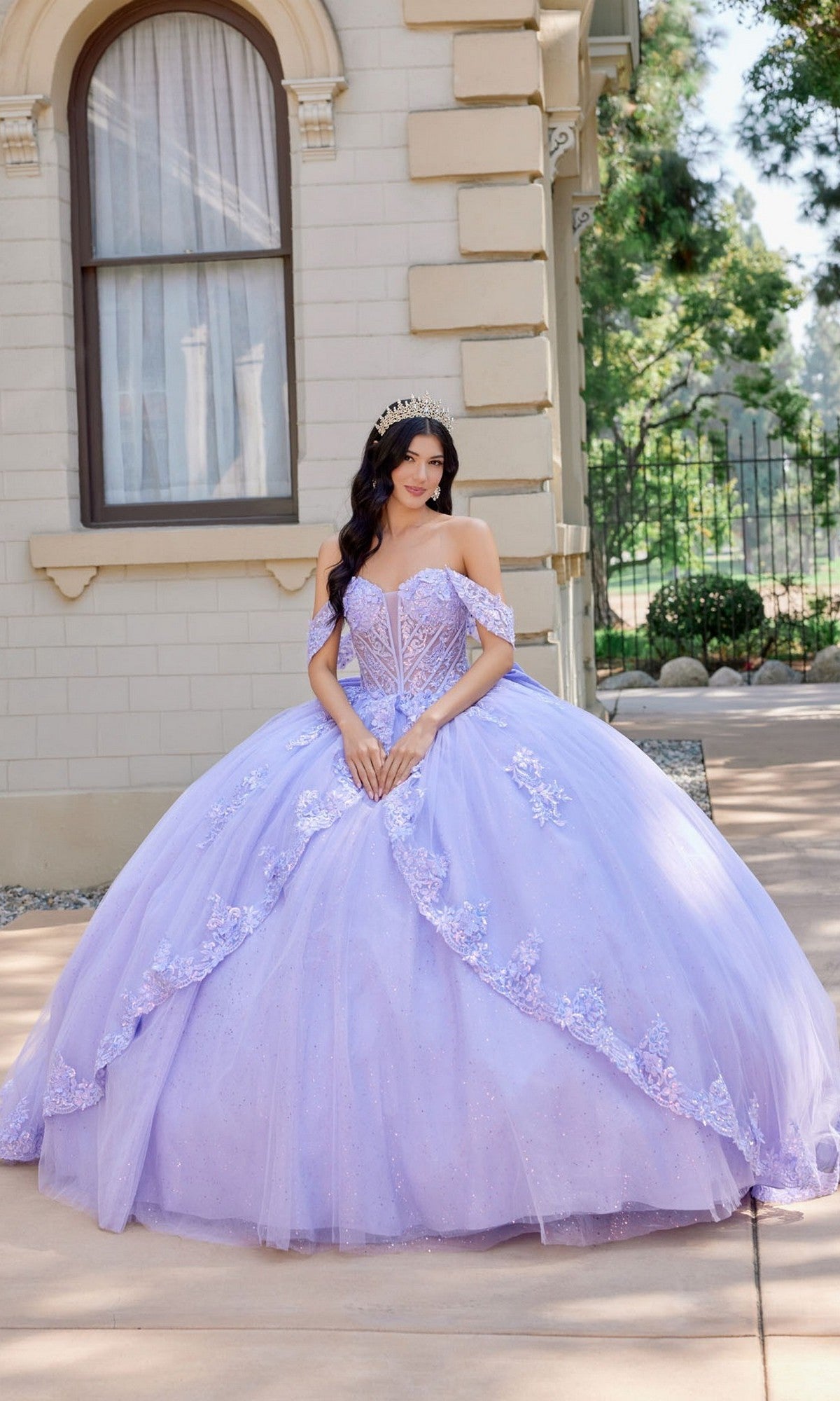 Wearing the Quinceanera Dress JT1507J by Juliet in lilac, a woman with a tiara stands outdoors near a beige building and lush greenery. The off-the-shoulder ballgown features delicate floral details and flowing tulle fabric.