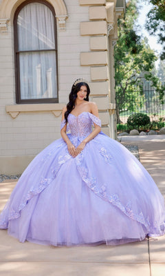 Wearing the Quinceanera Dress JT1507J by Juliet in lilac, a woman with a tiara stands outdoors near a beige building and lush greenery. The off-the-shoulder ballgown features delicate floral details and flowing tulle fabric.