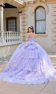 A woman wearing the Quinceanera Dress JT1507J By Juliet—a purple, off-the-shoulder gown adorned with floral details—and a tiara stands outside a building with white columns, facing away from the camera.