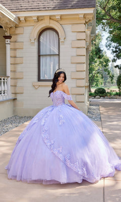 A young woman wearing the Quinceanera Dress JT1507J by Juliet and a tiara stands outside a cream-colored building, looking down and smiling.
