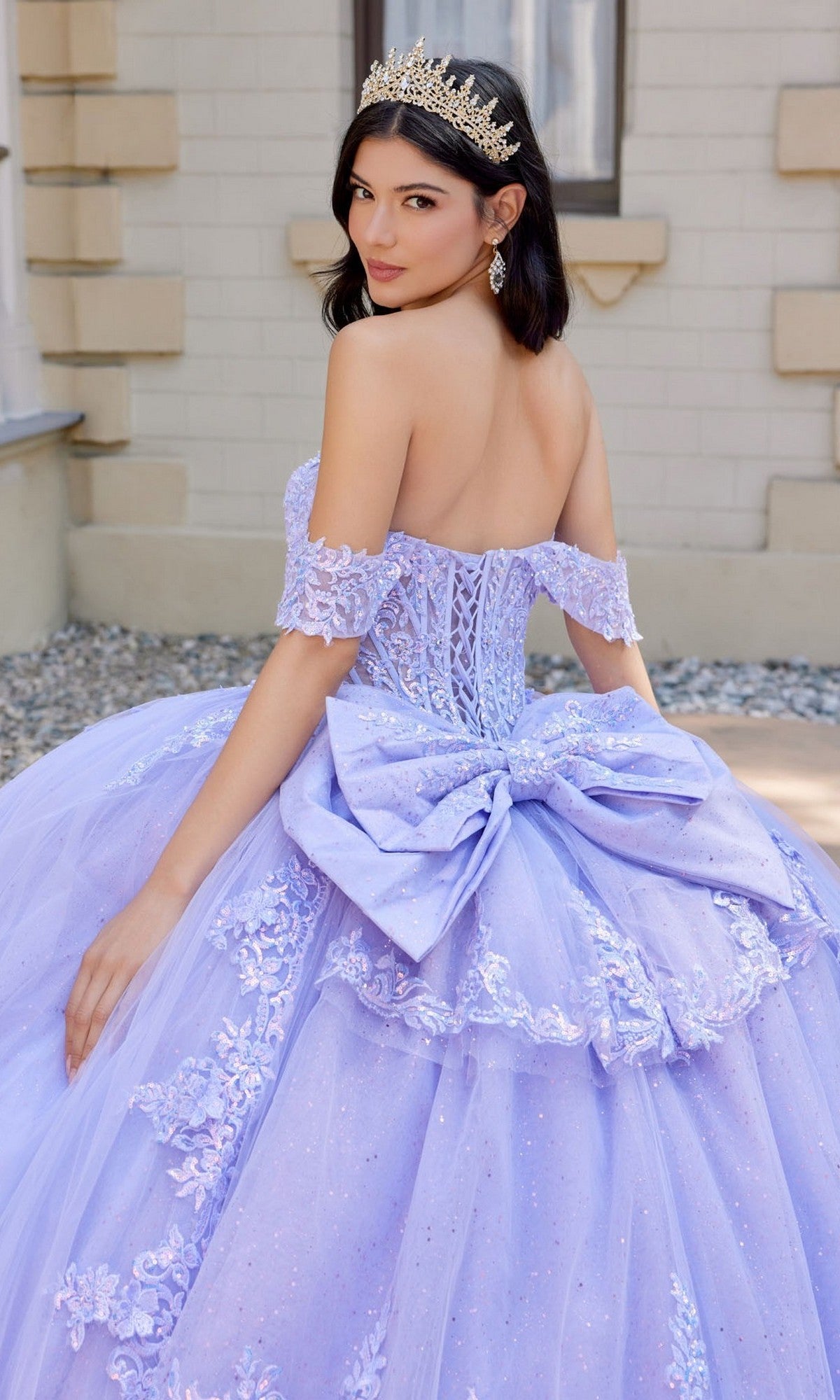 A woman stands outdoors adorned with a sparkling tiara and earrings, wearing the Quinceanera Dress JT1507J By Juliet—an elegant lavender off-the-shoulder tulle ballgown featuring a large bow and delicate lace details.