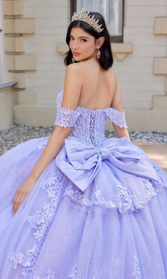 A woman stands outdoors adorned with a sparkling tiara and earrings, wearing the Quinceanera Dress JT1507J By Juliet—an elegant lavender off-the-shoulder tulle ballgown featuring a large bow and delicate lace details.