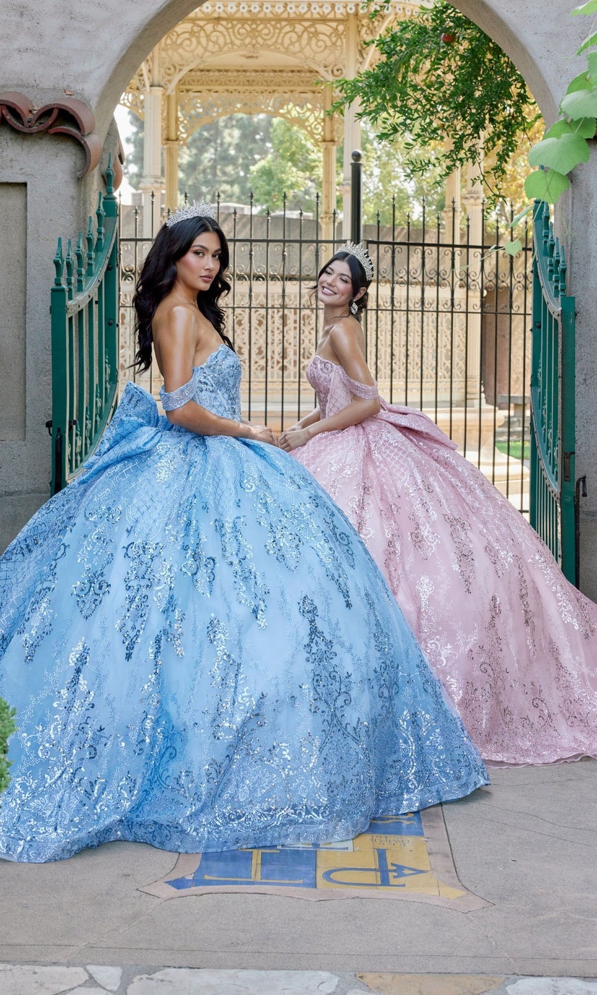 Two young women in elaborate, sparkling sequin ball gowns—one wearing the shimmering blue Quinceanera Dress JT1510H By Juliet with off-the-shoulder sleeves, and the other in a radiant pink gown—stand outdoors near a decorative gate and archway, posing gracefully for a formal portrait.