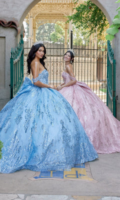 Two young women in elaborate, sparkling sequin ball gowns—one wearing the shimmering blue Quinceanera Dress JT1510H By Juliet with off-the-shoulder sleeves, and the other in a radiant pink gown—stand outdoors near a decorative gate and archway, posing gracefully for a formal portrait.