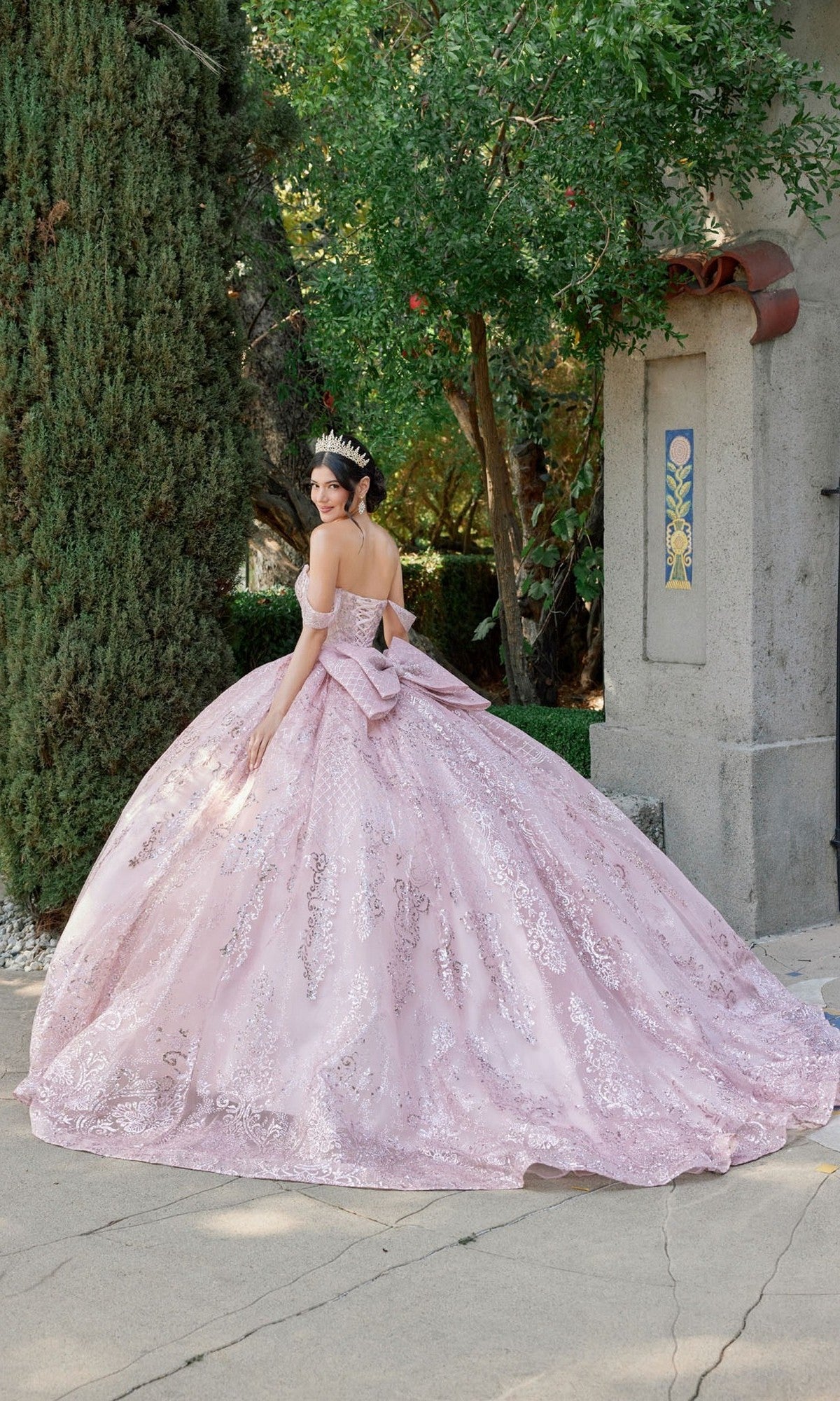 Wearing the blush Quinceanera Dress JT1510H by Juliet, a woman adorned with sparkling sequins and an elegant tiara stands outdoors beside tall trees and a stone structure, gazing back over her shoulder.