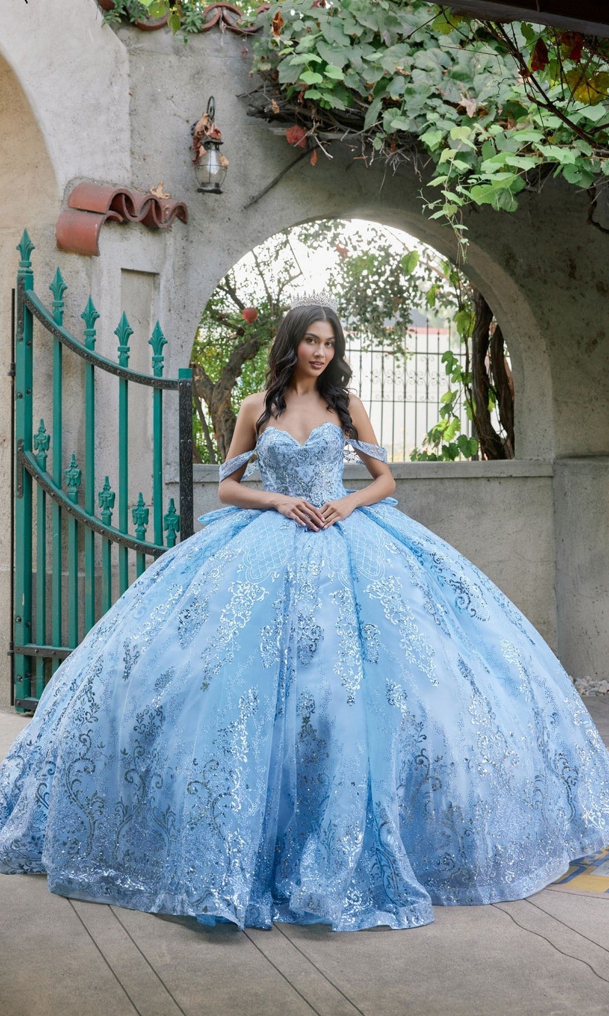 A woman in the Quinceanera Dress JT1510H By Juliet, a powder blue sequined ballgown, stands in front of a wrought iron gate and stone archway with her hands clasped.