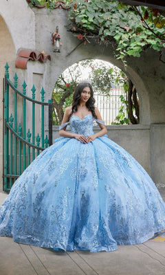 A woman in the Quinceanera Dress JT1510H By Juliet, a powder blue sequined ballgown, stands in front of a wrought iron gate and stone archway with her hands clasped.