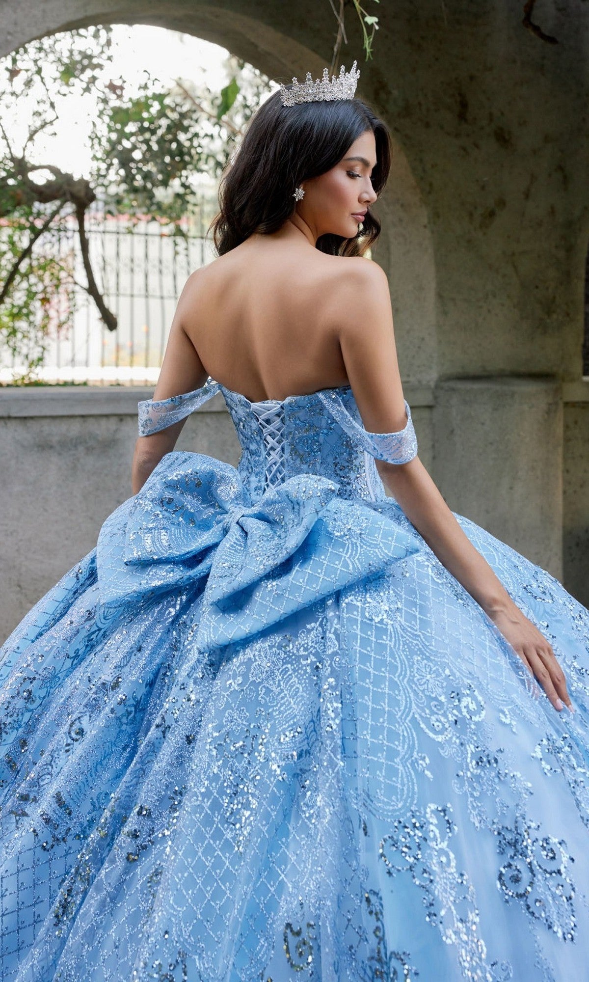A woman stands outdoors with her back mostly turned to the camera, wearing the Quinceanera Dress JT1510H By Juliet—a stunning off-the-shoulder ballgown embellished with sparkling sequins and a large bow—while a tiara rests elegantly atop her head.