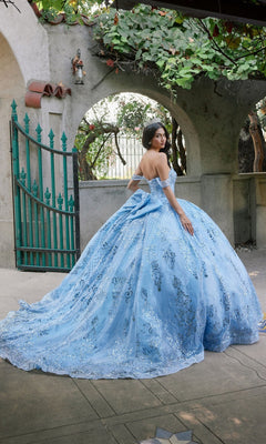 Wearing the Quinceanera Dress JT1510H By Juliet, a woman in an ornate blue ballgown adorned with sparkling sequins stands outdoors near a green gate, looking over her shoulder.