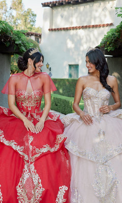 Two women wearing ornate ball gowns, including the stunning Quinceanera Dress JT1511H By Juliet with gold applique embroidery, sit next to each other outside, smiling and looking at each other.