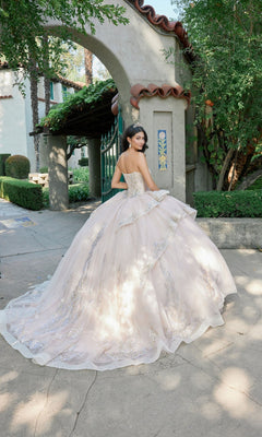 A young woman in the Quinceanera Dress JT1511H by Juliet, a strapless corset ballgown adorned with sequin gold applique, stands outdoors near an arched entryway amidst lush greenery and trees.