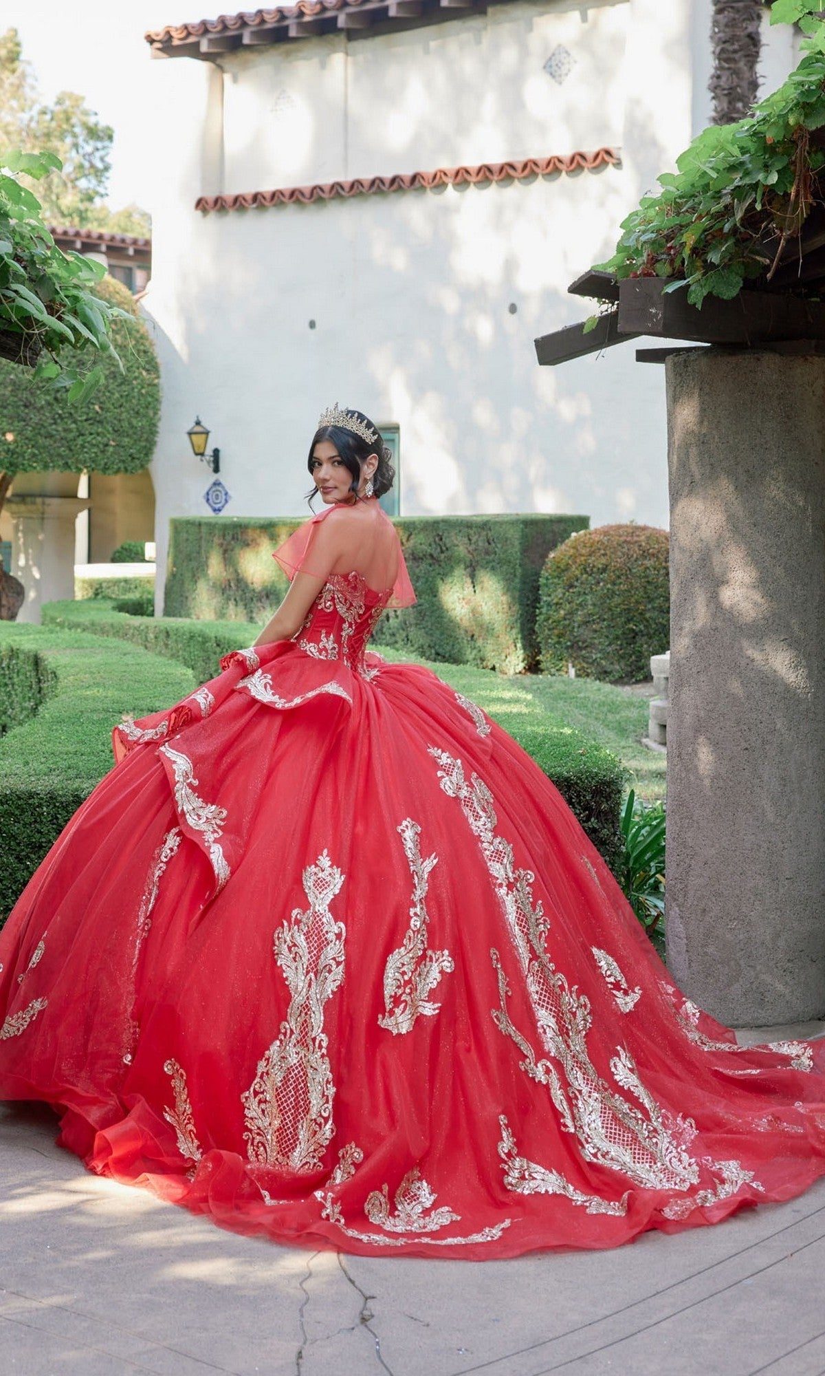 A young woman stands outdoors near a hedge, facing slightly away from the camera, wearing the Quinceanera Dress JT1511H By Juliet—a strapless ballgown featuring white embroidery.