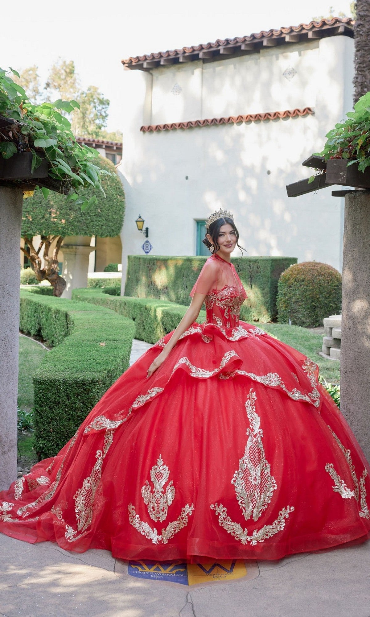 Wearing the Quinceanera Dress JT1511H By Juliet, a person stands outdoors in a garden, smiling at the camera in a red ballgown adorned with gold applique embroidery, paired with a tiara and gloves.