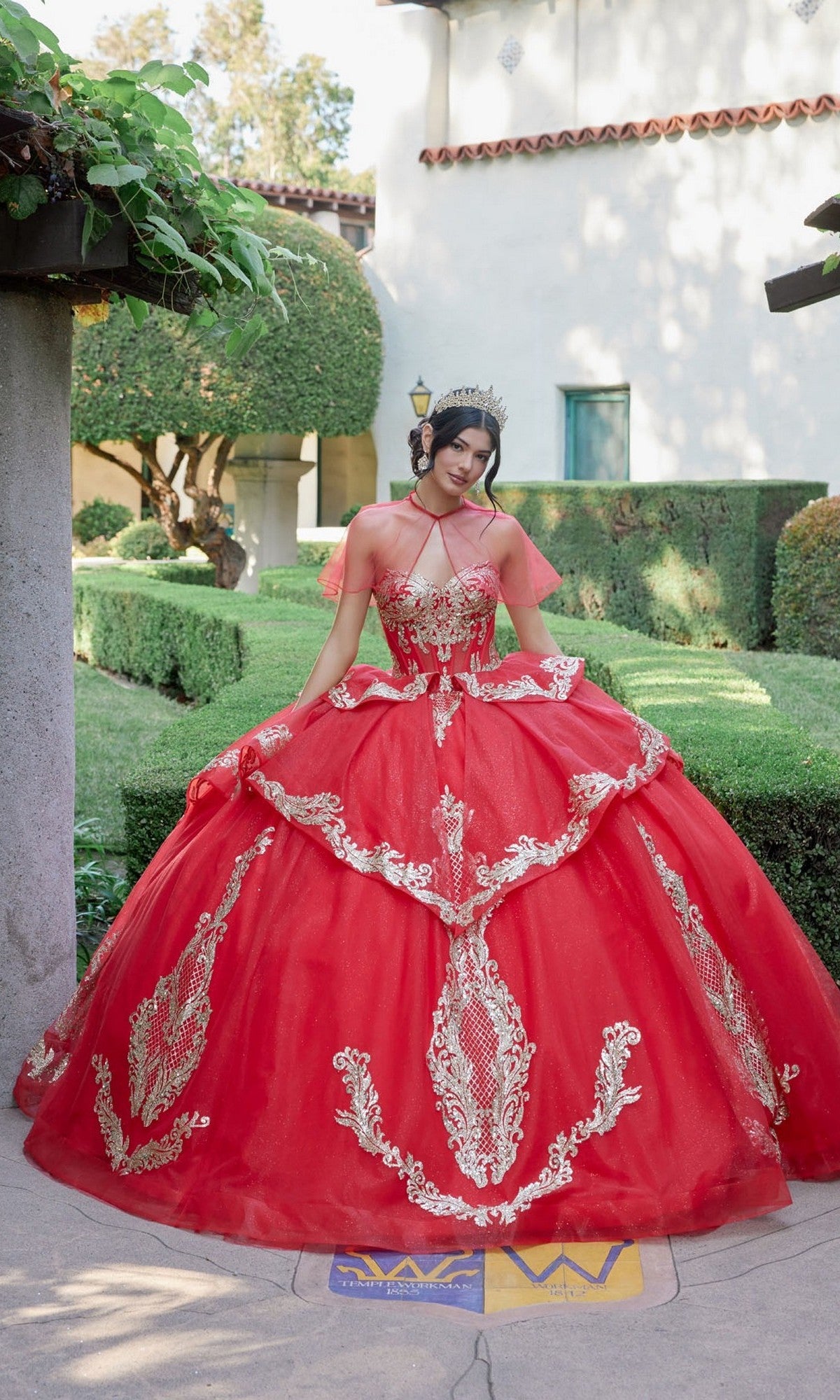 A young woman stands outdoors on a path, surrounded by manicured green bushes and a white building in the background, wearing the Quinceanera Dress JT1511H By Juliet—an ornate red and gold ballgown featuring intricate gold applique embroidery.