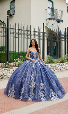 A young woman wearing the Quinceanera Dress JT1512H By Juliet in navy blue stands outside in front of a wrought iron fence and a building with green trim. The sparkling glitter ballgown skirt and elegant corset bodice of her gown add to her graceful presence.