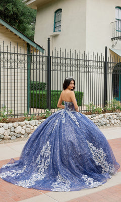 Wearing the Quinceanera Dress JT1512H by Juliet, a woman stands outside in front of a building and black iron fence. The strapless, blue and silver glitter ballgown features a corset bodice as she looks over her shoulder.