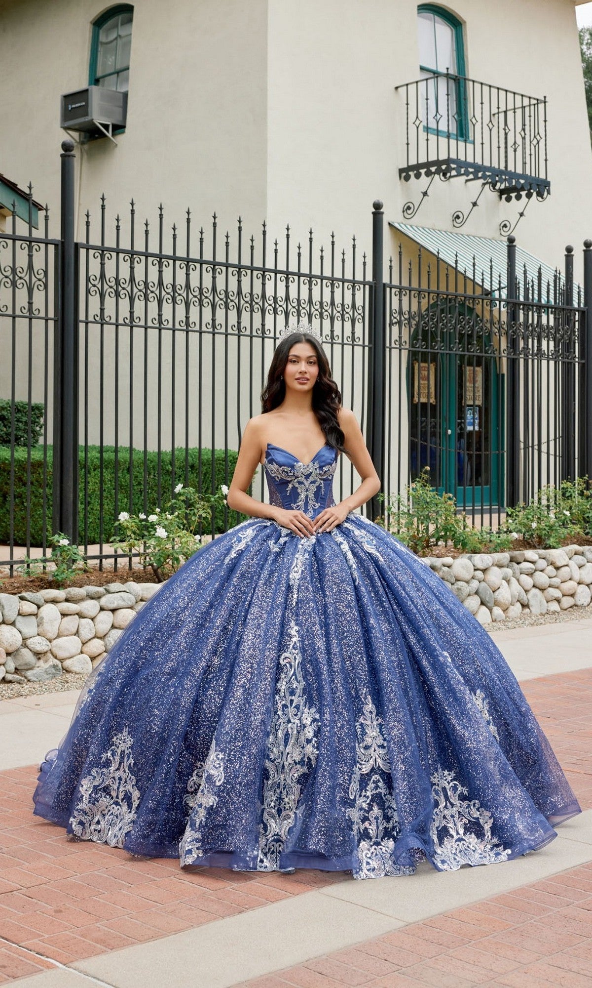 A young woman stands outdoors in front of a gated building, wearing the Quinceanera Dress JT1512H by Juliet—a stunning navy-blue gown featuring a strapless, corset bodice embellished with silver embroidery and a full, layered skirt.